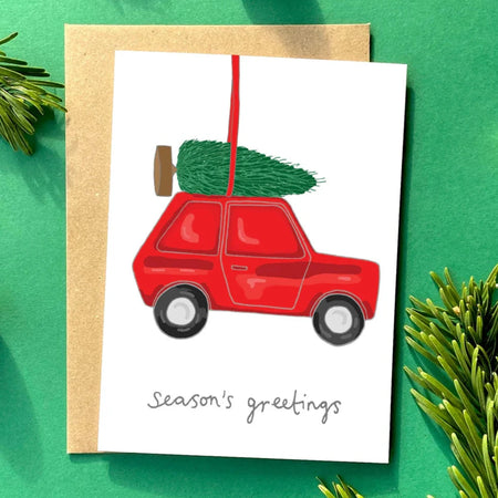 kitsch christmas card illustrated with a hanging tree decoration of a car with a christmas tree on the roof. and it reads seasons greetings.