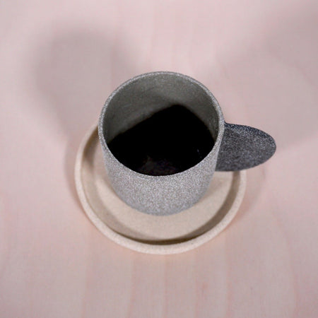 a grey espresso cup with a darker grey handle on a sand coloured saucer. sitting on a table.