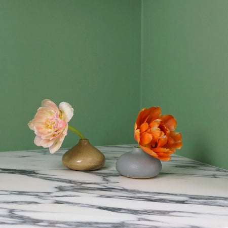 a duck egg blue bud vase with an orange flower and a oatmeal bud vase with a light pink flower on a marble table with a bright green background.