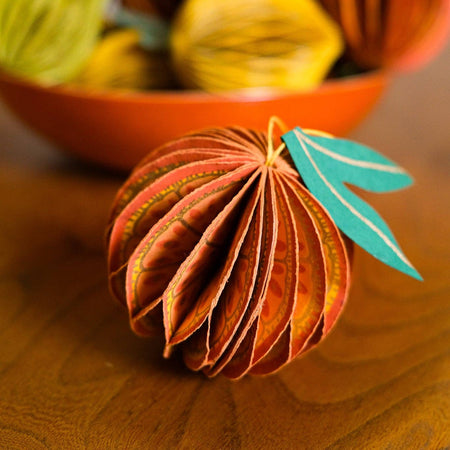 Decorative paper orange with leaf on a wooden surface, surrounded by colorful pumpkins in the background | East End Press | Shop Fodder