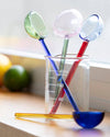 Three translucent glass measuring spoons in green, pink, and blue, with red handles, arranged in a clear 140ml beaker on a wooden surface. A yellow glass stirring rod and a blue spoon rest in front, with a softly blurred kitchen background containing lemons and limes.