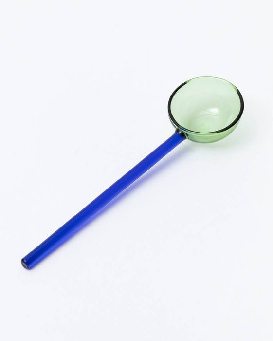 A blue glass drinking spoon with a light green bowl and rim, photographed diagonally on a clean white background.