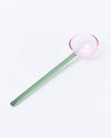 A long-handled glass spoon with a translucent green handle and a pale pink bowl, photographed on a clean white background.