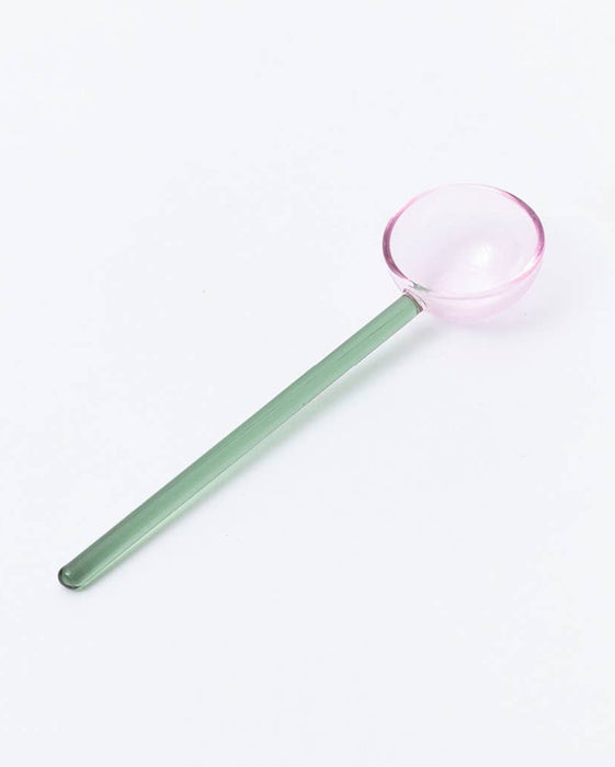 A long-handled glass spoon with a translucent green handle and a pale pink bowl, photographed on a clean white background.