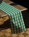 A green and white checkered table runner draped over a wooden slatted table on a wooden deck.