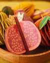 A close-up of a decorative pomegranate-shaped hanging ornament in pink and red with gold dotted details, displayed in a wooden bowl with other autumn-themed ornaments in the background.