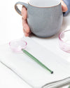 A gray ceramic mug with a handle is held by a hand over a white table setting. A translucent pink spoon with a green handle rests on a folded white napkin, and a small pink bowl containing white pearls and a green marble is on the right.