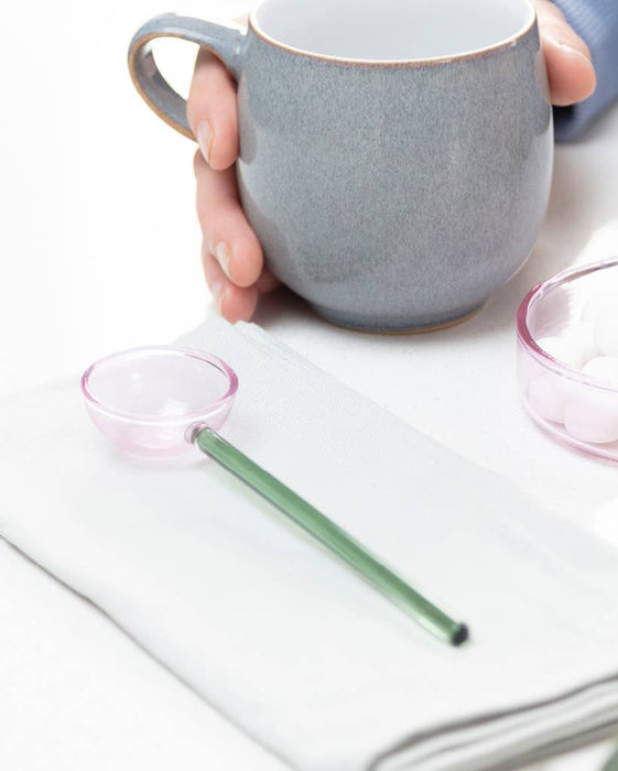 A gray ceramic mug with a handle is held by a hand over a white table setting. A translucent pink spoon with a green handle rests on a folded white napkin, and a small pink bowl containing white pearls and a green marble is on the right.