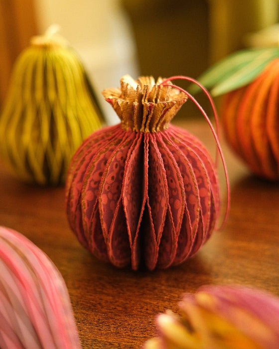 A close-up of a decorative paper pumpkin ornament in rust red with a textured pattern, standing on a wooden surface with other colorful paper gourds blurred in the background.