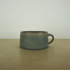handmade ceramic mug by aku ceramics. simple shape in a blue green glaze. on a wooden table with a white wall. 