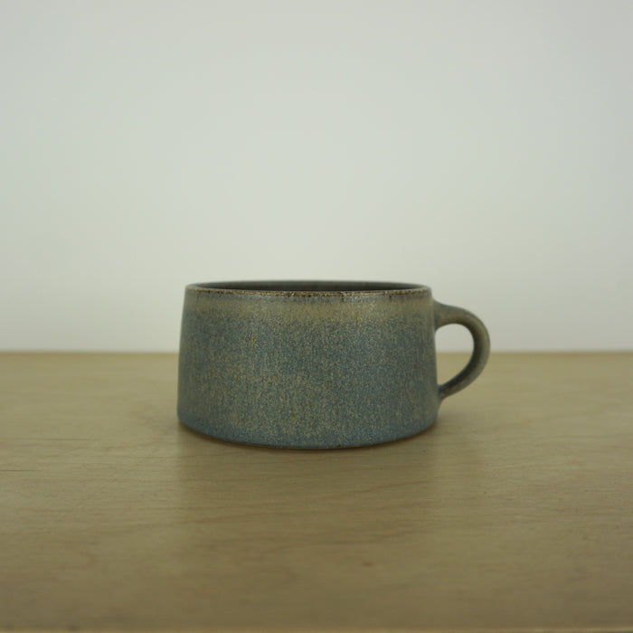 handmade ceramic mug by aku ceramics. simple shape in a blue green glaze. on a wooden table with a white wall. 