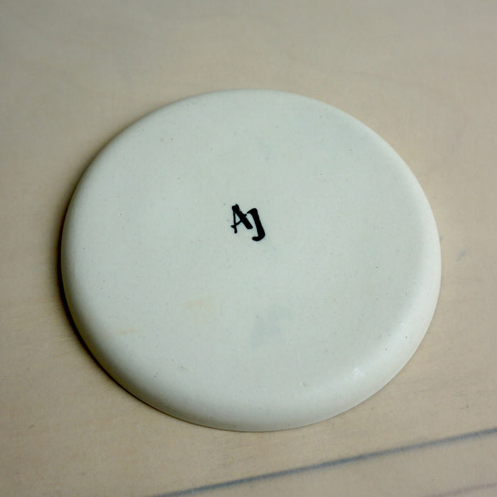 Round white object with 'AJ' marking on a beige background. Splatter Saucer | Anna Jones Ceramics | Shop Fodder