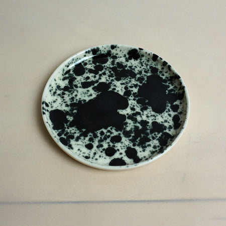 Speckled ceramic plate with black and white pattern on a beige background. Splatter Saucer | Anna Jones Ceramics | Shop Fodder