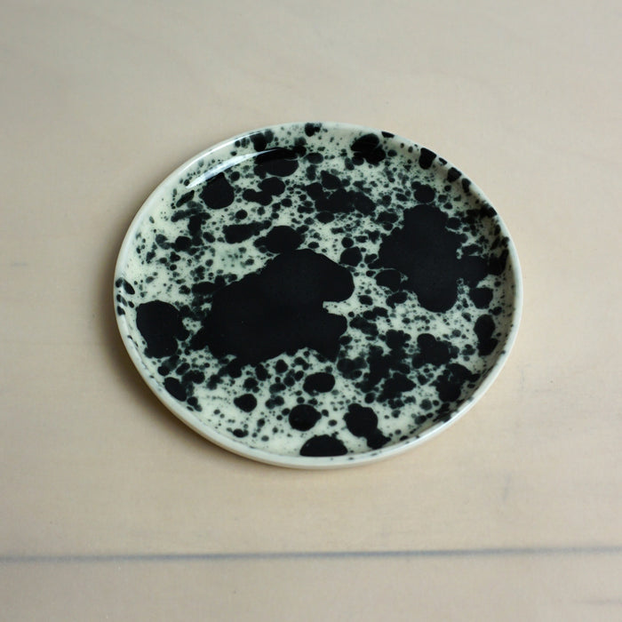Speckled ceramic plate with black and white pattern on a beige background. Splatter Saucer | Anna Jones Ceramics | Shop Fodder