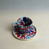 Ceramic cup and saucer with blue and red splatter design on a plain background. Splatter Saucer | Anna Jones Ceramics | Shop Fodder