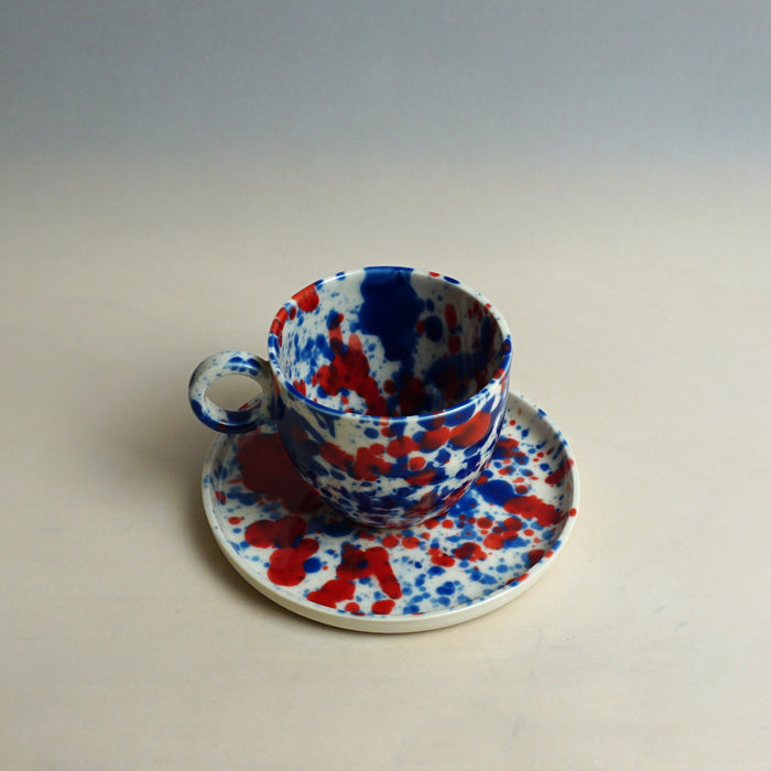 Ceramic cup and saucer with blue and red splatter design on a plain background. Splatter Saucer | Anna Jones Ceramics | Shop Fodder