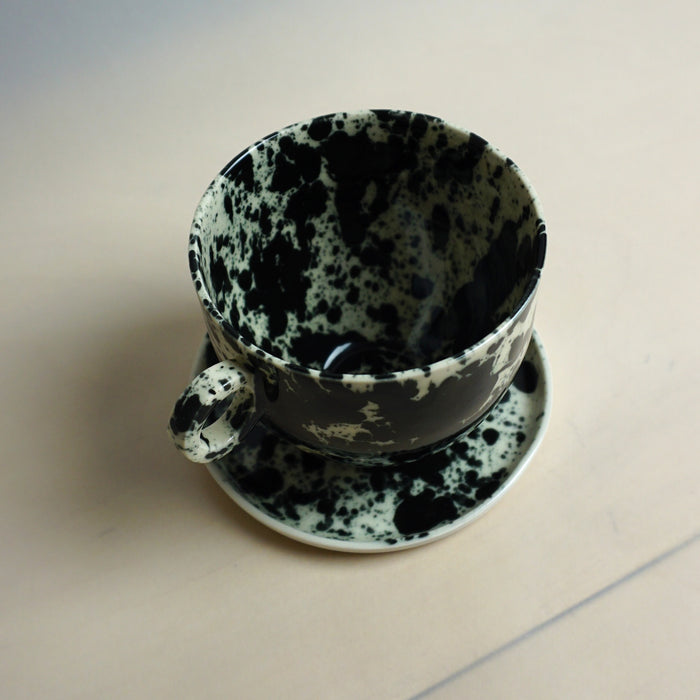 Speckled black and white ceramic cup and saucer on a plain background. Splatter Saucer | Anna Jones Ceramics | Shop Fodder