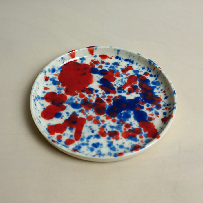 Round plate with red and blue splatter design on a beige background. Splatter Saucer | Anna Jones Ceramics | Shop Fodder