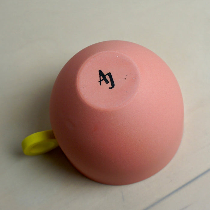 Pink mug with a yellow handle and 'AJ' makers mark on a light background. Syros Mug | Anna Jones Ceramics | Shop Fodder