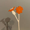 Two orange paper flowers on sticks against a gray background. Anna Wiscombe | Shop Fodder