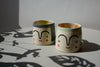 a green mug and a blue mug by beci callow both with faces hand painted on to them. 