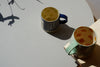 a green mug on a shelf and a blue mug on a table next to it. 