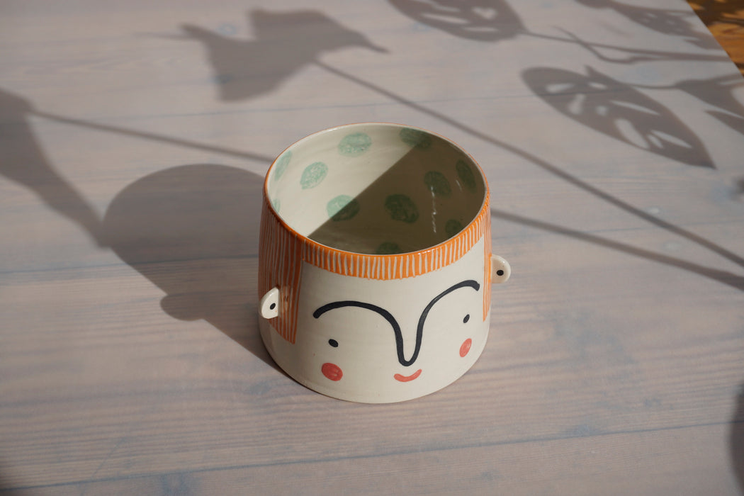 an orange planter with a face on the outside and green splodges on the inside. 
