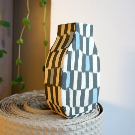 Checkerboard-patterned vase on a textured surface with a plant in the background