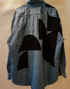 A blue denim shirt jacket with large black geometric patchwork panels on the back, shown against a plain beige background. Caroline Dickinson Shirt | Fodder