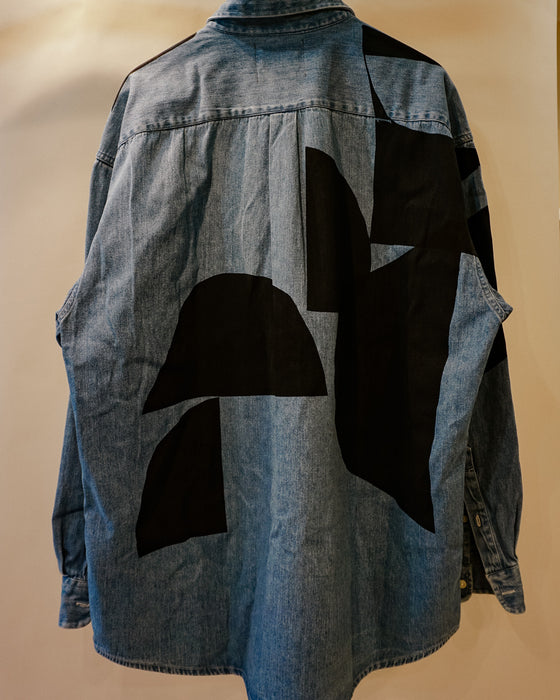 A blue denim shirt jacket with large black geometric patchwork panels on the back, shown against a plain beige background. Caroline Dickinson Shirt | Fodder
