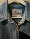 A close-up of a blue denim button-up shirt hanging on a white hanger, showing the neck label that reads Caroline Dickinson Brech. Caroline Dickinson Shirt | Fodder