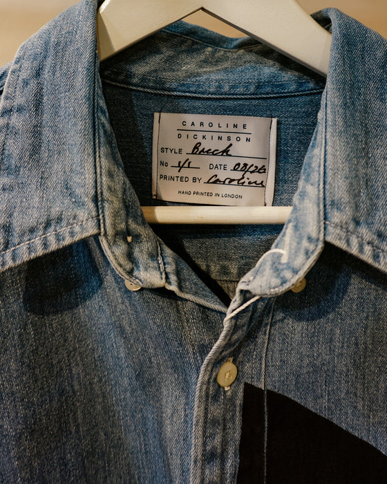 A close-up of a blue denim button-up shirt hanging on a white hanger, showing the neck label that reads Caroline Dickinson Brech. Caroline Dickinson Shirt | Fodder