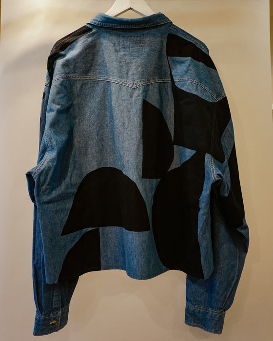 A blue denim jacket with large black abstract patchwork panels on the back, hanging on a white hanger against a plain beige wall. Caroline Dickinson Shirt | Fodder