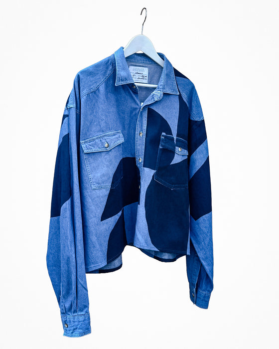 A blue patchwork denim shirt jacket hanging on a white hanger against a plain white background. The shirt features a mix of medium and dark blue denim panels, including a buttoned chest pocket and a large patch pocket on the front. Caroline Dickinson Shirt | Fodder