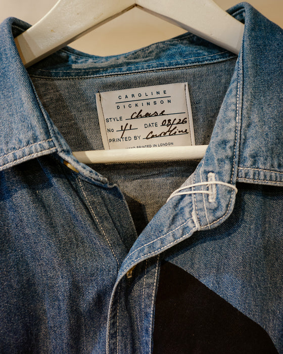 A close-up of a blue denim shirt jacket hanging on a white hanger, showing the collar, front button placket, and an interior neck label that reads Caroline Dickinson. Caroline Dickinson Shirt | Fodder