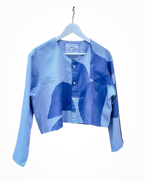 A cropped long-sleeve button-up shirt in a light blue denim-like fabric, featuring an abstract geometric pattern in various shades of blue. The shirt is displayed on a white hanger against a plain white background. Caroline Dickinson Shirt | Fodder