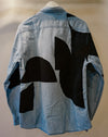 A light blue denim shirt jacket shown from the back, featuring large abstract black patchwork shapes over a light blue denim base, hanging on a white hanger against a plain beige wall. Caroline Dickinson Shirt | Fodder