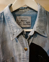 A light blue denim button-up shirt jacket with a pointed collar and a visible Carolina Dickinson brand label, hanging on a white hanger against a neutral background. Caroline Dickinson Shirt | Fodder
