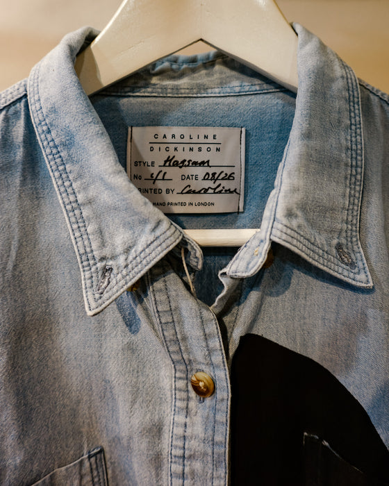 A light blue denim button-up shirt jacket with a pointed collar and a visible Carolina Dickinson brand label, hanging on a white hanger against a neutral background. Caroline Dickinson Shirt | Fodder