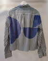 A long-sleeve button-up shirt hanging on a white hanger against a plain light background. The shirt features a black and white vertical stripe pattern with large blue abstract circular patches on the back. Caroline Dickinson Shirt | Fodder