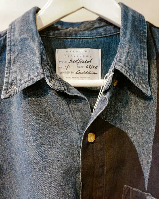 A close-up of a denim shirt jacket hanging on a white hanger, showing a collar, front button placket, and a sewn-in label that reads Caroline Dickinson Style Redfield No. 1. Caroline Dickinson Shirt | Fodder