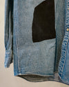 A close-up of a light blue denim button-up shirt with a large black fabric patch on the front, shown hanging against a plain light background. Caroline Dickinson Shirt | Fodder