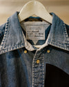 A close-up of a denim jacket hanging on a white hanger, showing a blue denim collar, striped inner shirt, brass buttons, and a visible Carolina Dickinson neck label. Caroline Dickinson Shirt | Fodder