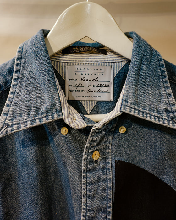A close-up of a denim jacket hanging on a white hanger, showing a blue denim collar, striped inner shirt, brass buttons, and a visible Carolina Dickinson neck label. Caroline Dickinson Shirt | Fodder