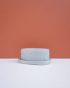 A small round gray textured planter pot with a matching saucer, set against a split background of white and terracotta orange. Brutes Ceramics | Hope Collection | Shop Fodder