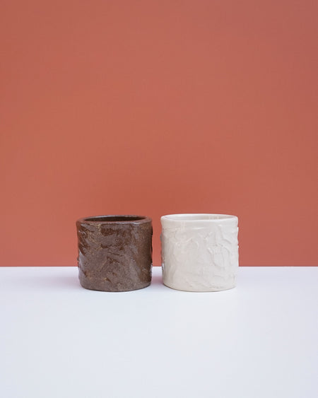 Two textured ceramic planters, one dark brown and one off-white, displayed side-by-side on a white surface against a terracotta-colored background. Brutes Ceramics | Hope Collection | Shop Fodder