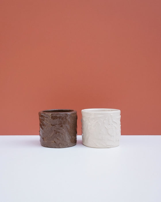 Two textured ceramic planters, one dark brown and one off-white, displayed side-by-side on a white surface against a terracotta-colored background. Brutes Ceramics | Hope Collection | Shop Fodder