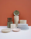 A collection of textured ceramic planters and shallow dishes in earthy tones, including beige, gray, green, and pink, arranged on a white surface against a terracotta background. Brutes Ceramics | Hope Collection | Shop Fodder