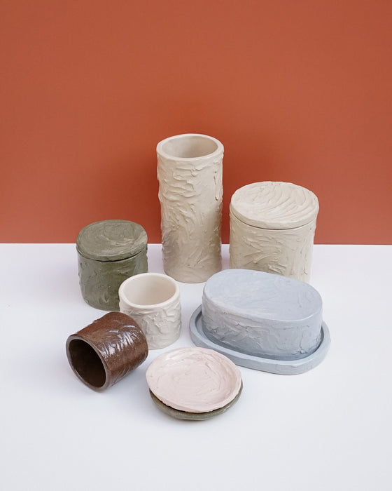 A set of six textured ceramic storage containers and vessels in various earthy tones, including beige, gray, brown, and pink, arranged on a white surface against a terracotta background.  Brutes Ceramics | Hope Collection | Shop Fodder