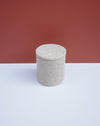 A small cylindrical lidded container with a textured, hand-painted finish in a light warm gray-beige color, set against a split background of white and terracotta red. Brutes Ceramics | Hope Collection | Shop Fodder